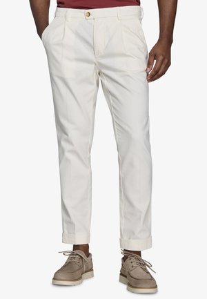 Mężczyzna ubrany w kremowe spodnie z mankietami, brązowe sznurowane buty i bordo koszulę, stojący z częściowo wsuniętymi dłońmi do kieszeni.