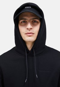 Felpa nera con cappuccio foderato e cordini, con il logo "Peak Performance" stampato in modo tonale e discreto. Cappello scuro con logo.