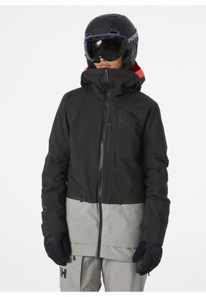 Zwarte en grijze skijas met een rits aan de voorkant, capuchon en elastische manchetten; gestructureerd ondergedeelte; handschoenen en helm gedragen door het model.