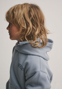 Halli dressipluus paelaga, valmistatud pehmest kangast. Omab tikitud logot ja kihilist särki all, millel on sinised triibud.