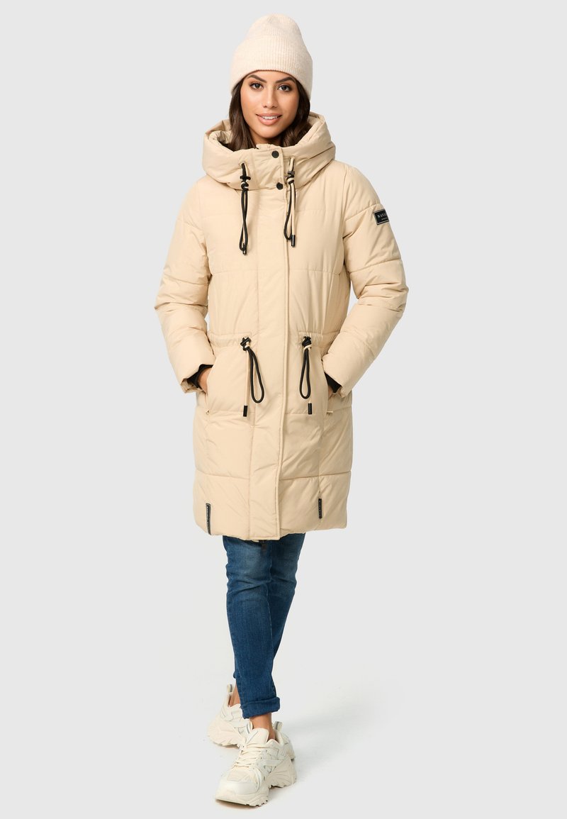 Beige pufferjack met een capuchon, verstelbare trekkoorden, zijzakken en zwarte accenten. Draagt over een blauwe spijkerbroek en witte sneakers.