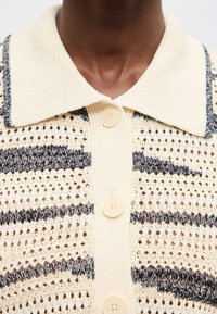 Κοντινή λήψη κρεμ πλεκτής πόλο μπλούζας με μαύρες οριζόντιες ρίγες και κρεμ κουμπιά στον λαιμό και στο επάνω μέρος του στήθους ενός σκούρου δέρματος ατόμου.