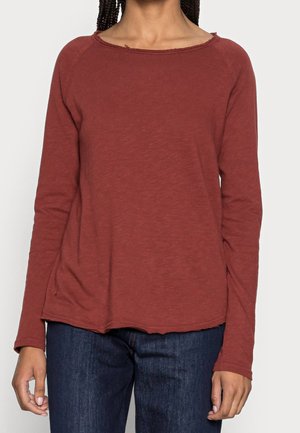Person iført en langærmet, rustfarvet skjorte med rund halsudskæring og mørkeblå jeans mod en ensfarvet lys baggrund.