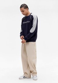 Felpa blu navy con maniche grigie, con un logo sul davanti, abbinata a pantaloni beige a gamba larga e sneakers bianche.