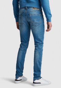 Muž v modrých taperovaných džínách, bílých teniskách a modrém tričku s dlouhým rukávem, stojící zády k fotoaparátu na bílém pozadí.