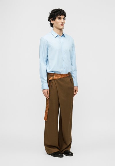 Jeune homme aux cheveux bouclés portant une chemise bleu clair à boutons, un pantalon large marron et une longue ceinture en cuir marron clair portée flottante.