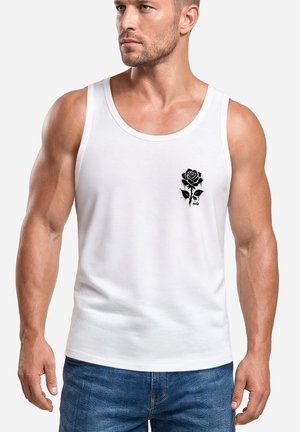 Man wearing a white sleeveless tank top with a small black rose graphic on the left chest and blue jeans, looking to the side.