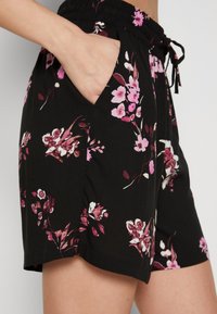 Main dans la poche d’un short noir avec un motif floral rose et blanc, taille élastique et cordon de serrage.