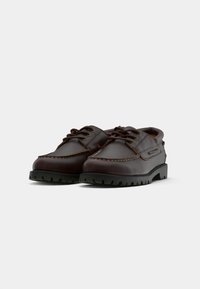 Par de sapatos de pele castanho-escura com atacadores, solas em borracha preta grossa e costura detalhada sobre um fundo branco.