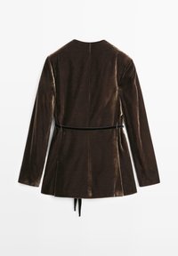 Brun fløjlsblazer med lange ærmer, en høj krave og en bælte i taljen. Har sideslidser og en glat, skinnende tekstur.