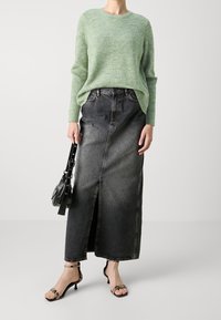 Helles grünes Strickpullover, dunkelgraer Jeans-Maxi-Rock mit einem Schlitz vorn, schwarze Handtasche und sandalen mit hohen Absätzen und Schlangenleder-Muster.