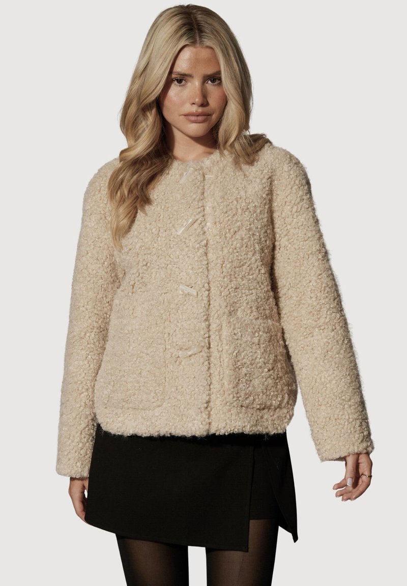 Flødefarvet fleecejakke med en tekstureret, fluffy overflade. Har rund hals, tre frontknapper og to sidelommer.
