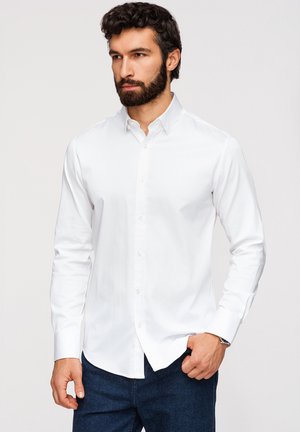 Homme aux cheveux foncés et à la barbe, portant une chemise blanche boutonnée et un pantalon bleu foncé, debout, une main dans la poche.