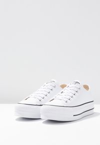 Zapatillas de lona blancas con una textura suave, diseño de corte bajo, detalle de franjas negras y ojales de metal para los cordones. Suelas de goma.