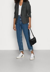 Grau strukturierter Blazer über einem weißen V-Ausschnitt-Shirt, kombiniert mit hochgekrempelten blauen Jeans und weißen Sneakers. Eine kleine schwarze Handtasche rundet den Look ab.