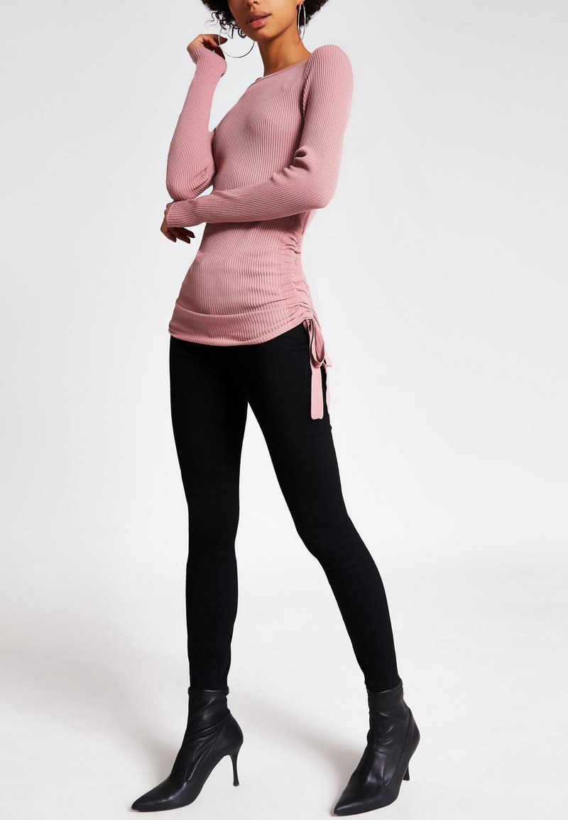 Langärmliges, geripptes Oberteil in sanftem Rosa, mit seitlichen Raffung und Bändern. Kombiniert mit figurbetonten schwarzen Hosen und spitzen schwarzen Ankle Boots.