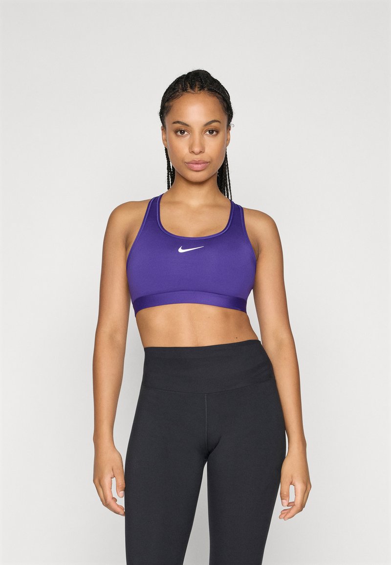 Femme avec des cheveux tressés portant un soutien-gorge de sport Nike violet et un legging noir taille haute sur un fond clair.