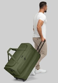 Uomo che indossa una maglietta bianca e pantaloni beige sta trascinando una grande borsa a rotelle color verde oliva su uno sfondo grigio.