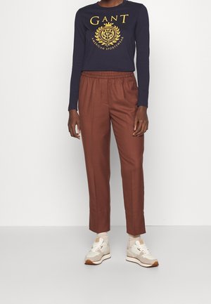 Persona che indossa un top a maniche lunghe blu navy con il logo GANT giallo, pantaloni marroni con cintura elastica, calzini bianchi e sneakers beige in piedi davanti a uno sfondo bianco.