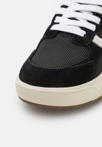 Sorte semsket og mesh joggesko med hvite snørebånd, en teksturert kremfarget mellomsåle og en brun gummisåle. Har et moderne atletisk design.
