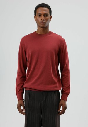 Rode trui met lange mouwen in een zachte brei met een ronde hals, gedragen met zwarte pantalon met krijtstreep. De pasvorm is ruim en de textuur is glad.