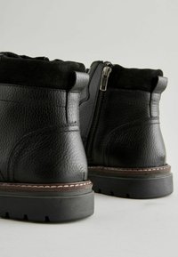 Botas de tobillo de cuero negro con un acabado texturizado, puntera redondeada, cremallera lateral y collarín acolchado. Presenta un cosido en marrón en la suela que contrasta.
