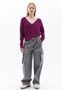 Top magenta con scollatura a V e maniche lunghe, abbinato a pantaloni cargo grigi con tasche laterali e vestibilità ampia, su uno sfondo neutro.