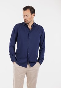 Mann med skjegg iført en marineblå knappeskjorte og beige bukser, hendene i lommene, ser ned og til siden mot en hvit bakgrunn.