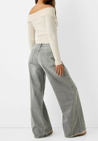 Krémový svetr s copánkovým úpletem a odhalenými rameny, spojený s vysokým pasem a širokými šedými džíny s volným střihem a dvěma zadními kapsami.