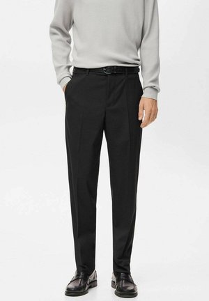 Hombre con pantalones negros a medida con bolsillos laterales, cinturón negro, suéter gris de manga larga con textura acanalada y mocasines de cuero negro sobre fondo blanco.