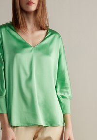Blouse en satin vert avec un col en V, des manches trois-quarts et une coupe décontractée. Texture lisse avec un léger éclat. Design sobre, accents minimalistes.