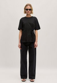 Zwarte katoenen T-shirt met korte mouwen en een zijstrikdetail, gecombineerd met loszittende zwarte jeans en platte sandalen. Rond de halslijn en een gladde textuur.