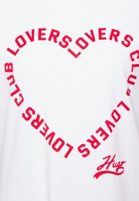 Hvid T-shirt med et markant rødt hjerte-design lavet af gentaget tekst: "LOVERS CLUB" i fede bogstaver, med "Hugo" i kursiv nederst.