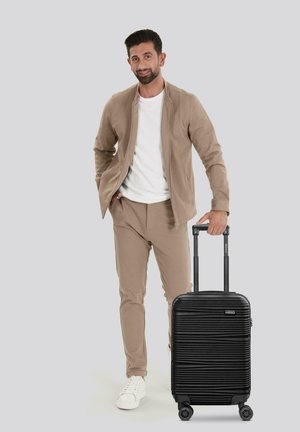 Zwarte harde koffer met een gestructureerd ribbelontwerp, dubbele wielen en een telescopische handgreep, geplaatst naast een persoon in beige kleding.