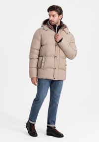 Beige puffjacka med huva, framfickor och kviltad design. Bärs över en svart zip-up tröja och kombineras med blå jeans och svarta stövlar.