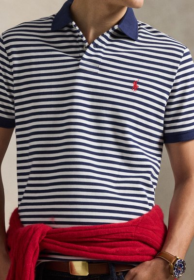 Άνδρας που φοράει μπλε ναυτικό και λευκό ριγέ πόλο πουκάμισο με κόκκινο πουλόβερ δεμένο γύρω από τη μέση και καφέ δερμάτινο ρολόι στον καρπό.