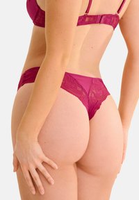 Magenta kantelaars met transparante panelen, bloemaccenten en een delicate tailleband. Heeft een V-vormige achterkant voor minimale bedekking.