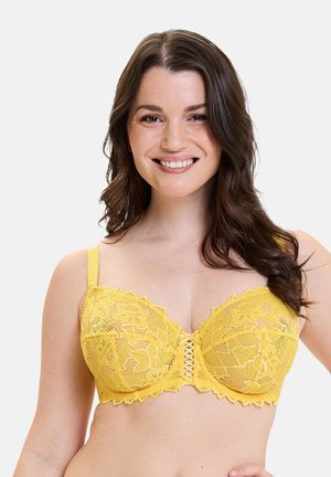 Glunderende vrouw met lang donker haar, dragen van een gele kanten bh met bloemmotieven tegen een witte achtergrond.