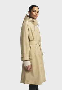 Kvinde iført en beige regnfrakke med bælte, hætte og strikkede manchetter, stående sidelæns mod en ensfarvet lysegrå baggrund.