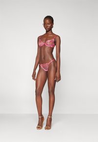Lingeri-sæt i lys pink blonde med blomster mønstre, der omfatter en balconette-bh og matchende g-streng, suppleret med guldhæle.