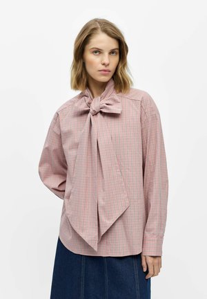 Femme portant un chemisier à carreaux rose avec un nœud oversize au col et une jupe bleu foncé, debout devant un fond blanc uni.