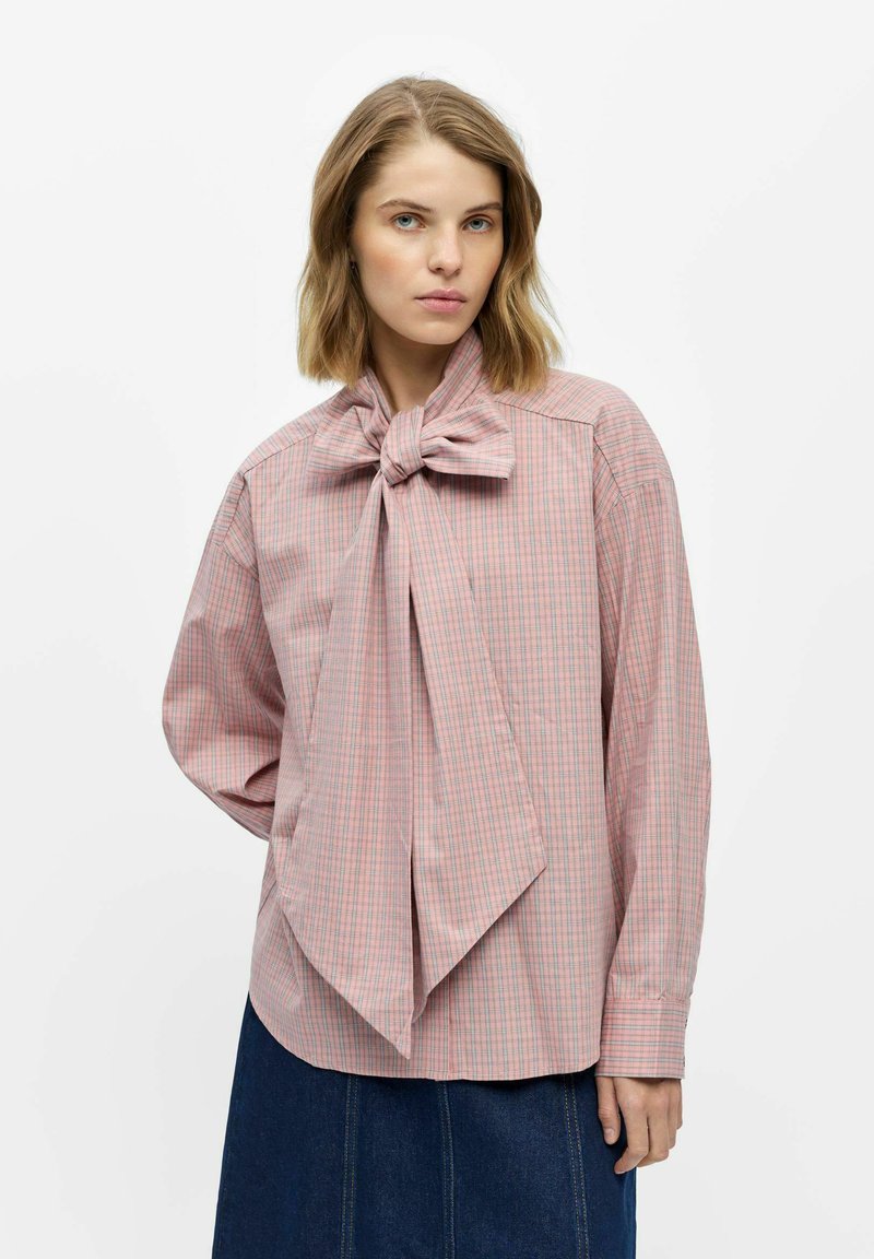 Femme portant un chemisier à carreaux rose avec un nœud oversize au col et une jupe bleu foncé, debout devant un fond blanc uni.
