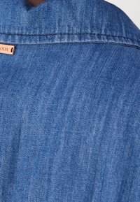Blaue Jeanshemd mit Kragen, glatter Textur und sichtbaren Nähdetails. Kleines Markenlabel an der Kragennaht.