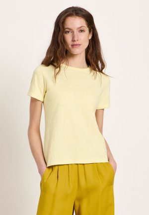 Frau mit langen, welligen Haaren, die ein blassgelbes kurzärmeliges T-Shirt und senfgelbe plissierte Hosen trägt, die Hände in den Taschen, neutraler Hintergrund.