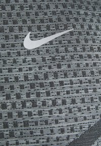 Zbliżenie szarego materiału z czarnym teksturowanym wzorem oraz białym logo Nike w kształcie swoosh, umieszczonym na środku materiału.