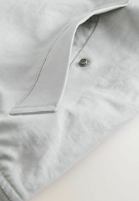 Camisa de tela gris claro con cuello, cierre de botones y superficie texturizada con detalles de costura visibles.