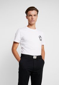 Camiseta de algodón blanca con cuello redondo y mangas cortas, con un diseño gráfico negro en el lado izquierdo del pecho, combinada con pantalones negros y un cinturón con hebilla plateada.