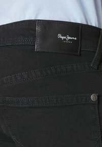 Crne traperice od denima s kožnom etiketom na kojoj piše "Pepe Jeans London." Sadrži metalne hardverske dijelove i suptilne detalje šavova.