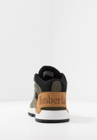Zapatilla verde y negra con una parte superior de ante texturizado, paneles de malla y una suela de goma. Presenta un parche de cuero marrón en el talón con el texto "Timberland" embossado.