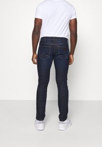 Jeans de mezclilla azul oscuro con un corte ajustado, que presentan bolsillos traseros con solapas puntiagudas, detalles de costura y un parche de cuero en la cintura.